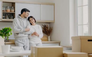 A female stood leaning on a male against a kitchen worktop looking at moving boxes.