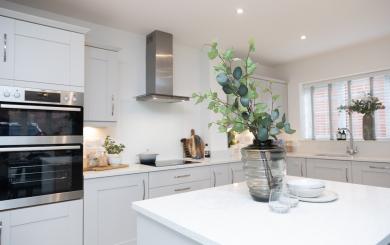 Interior of a clean and tidy white kitchen with cream cabinets. In the foreground is a kitchen island on which is a vase with foliage in and some crockery next to it