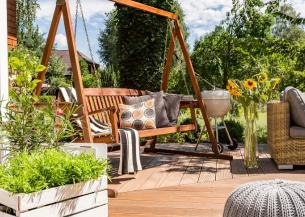 A wooden decking area with a swing seat, sunflowers, a barbecue and more plants 